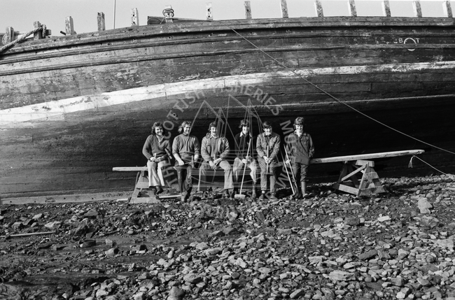 Group portrait of men seated in front of boat in